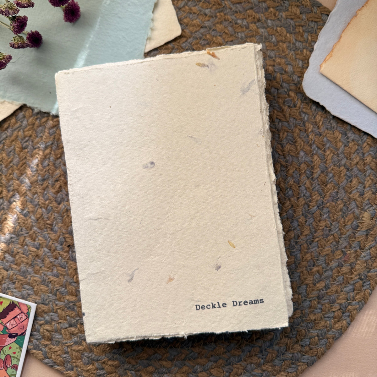 A handmade paper notebook with rough, unbleached edges and the text 'Deckle Dreams' printed in black at the bottom right, resting on a brown woven placemat.