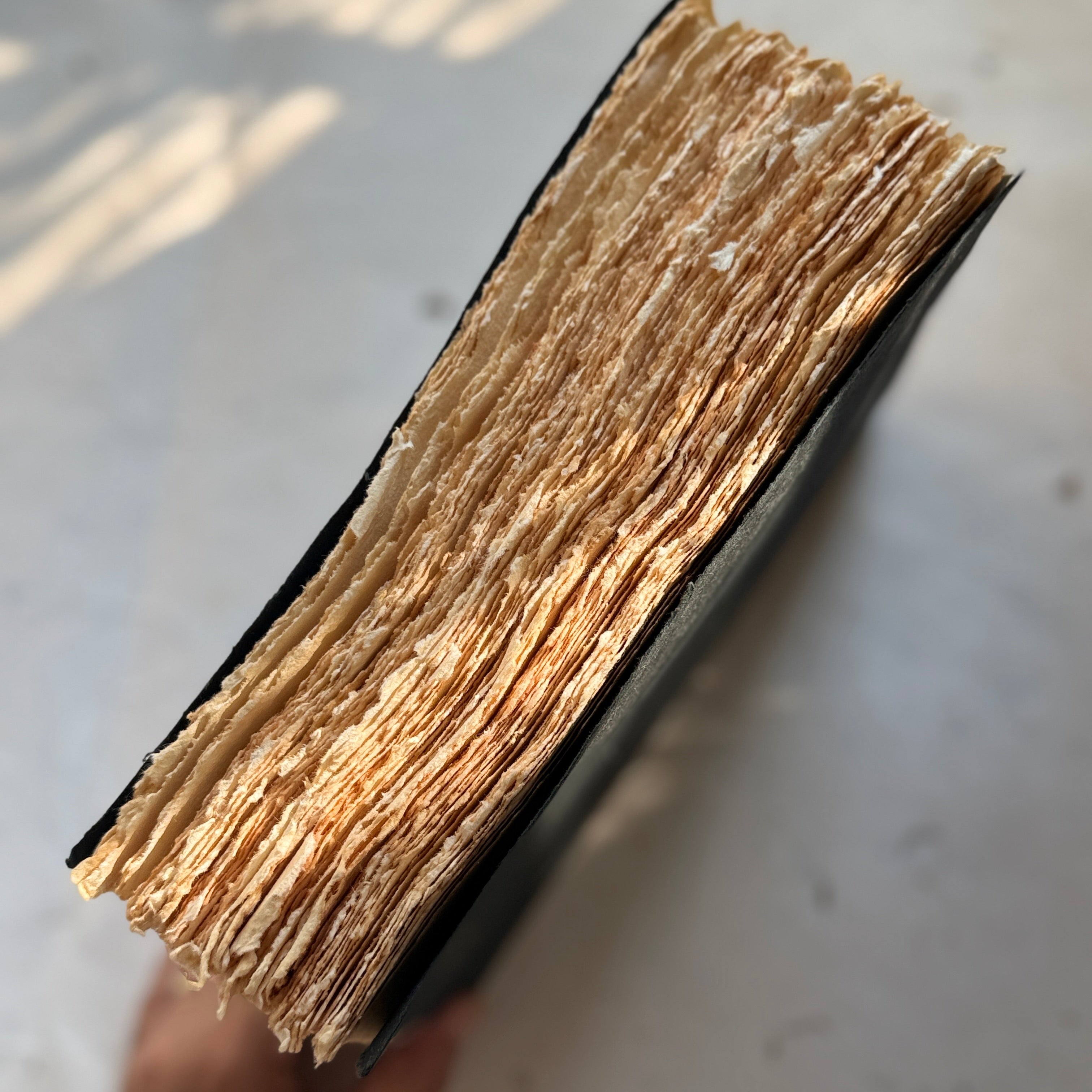 A hand holding the edge of a thick, vintage-style book or journal, showing a dense stack of aged, yellowed paper pages against a light gray background with soft shadows.