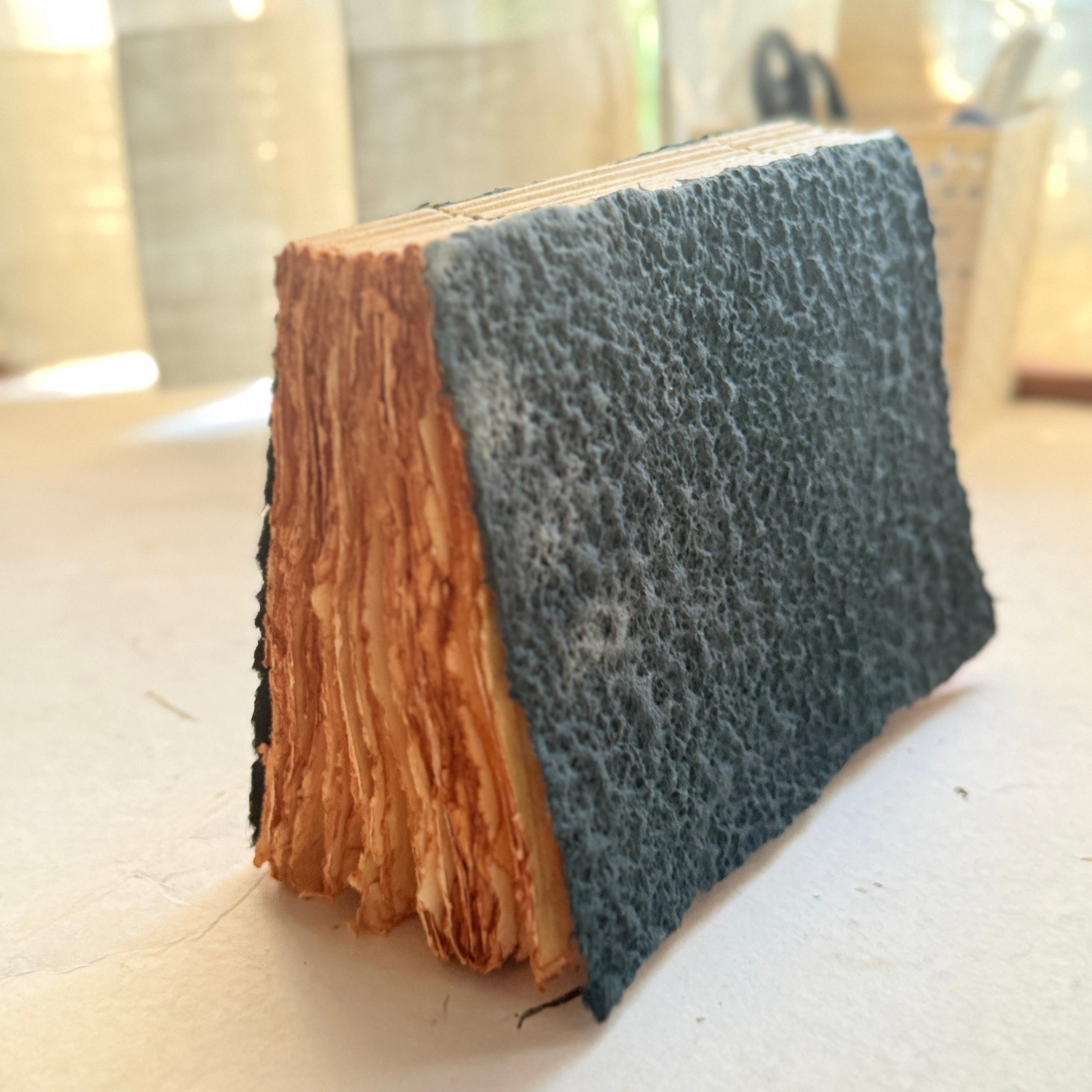 A handmade leather-bound book or journal standing upright on a light surface, with textured dark blue-gray leather on the cover and visible aged tan paper pages along the spine.
